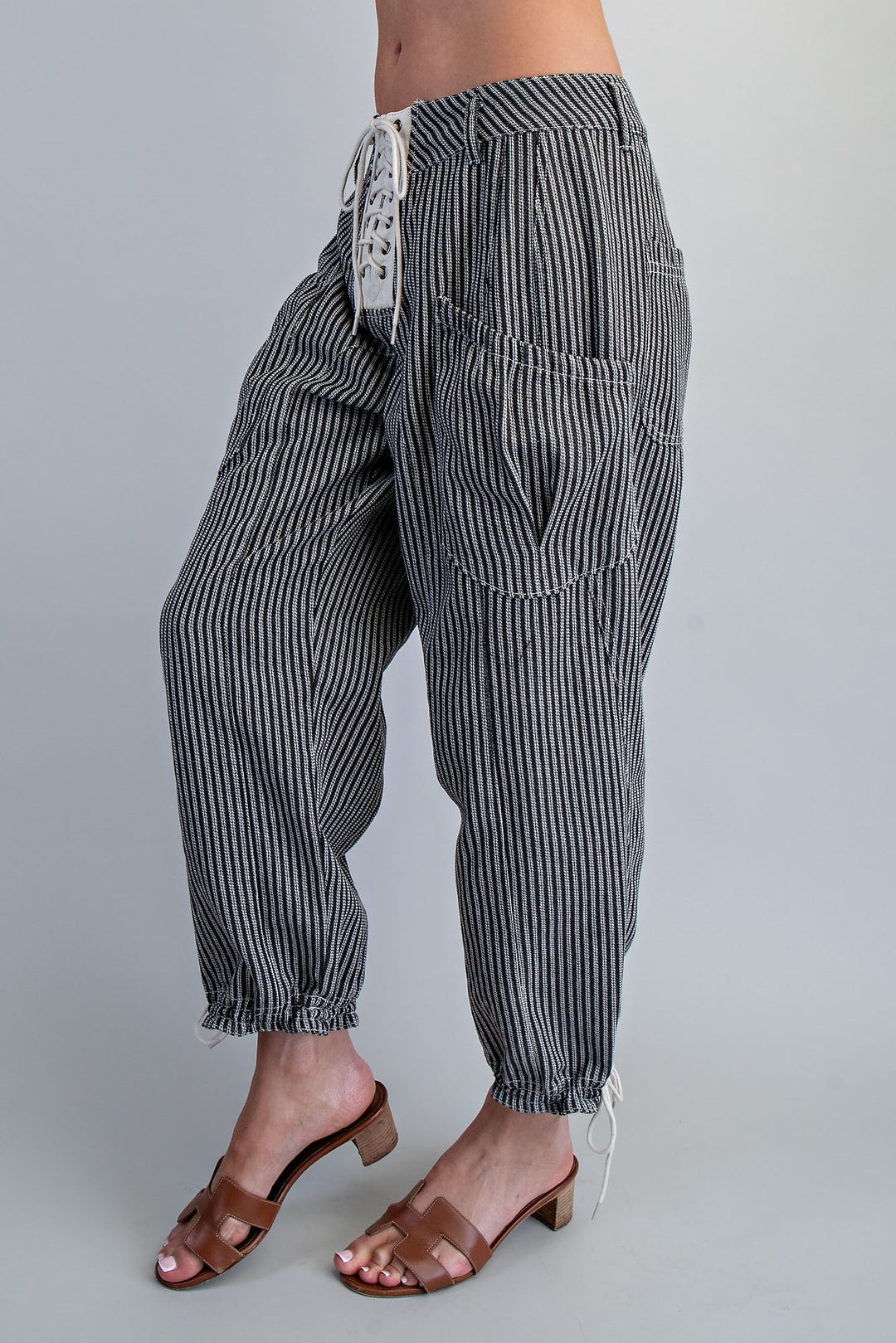 A person wearing loose black-and-white vertically striped drawstring pants with large front patch pockets and elastic cuffed ankles, paired with brown heeled slide sandals against a light gray studio background.