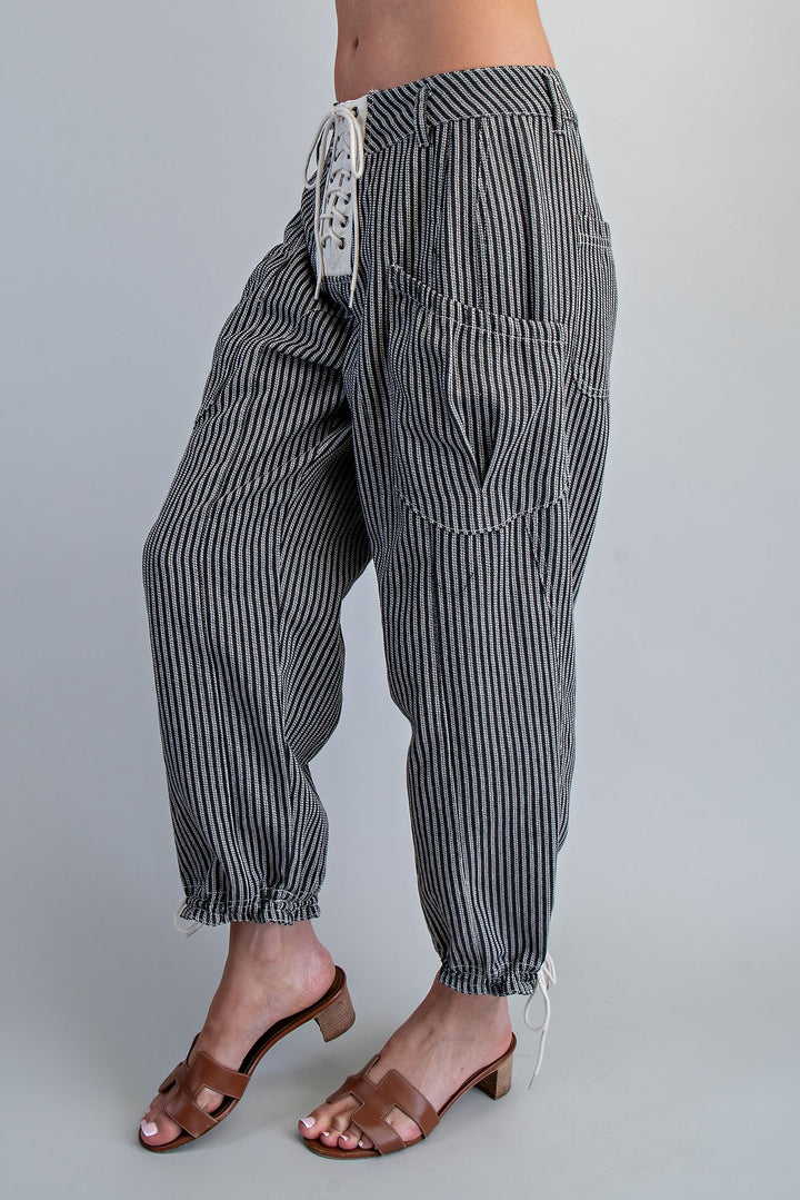 A person wearing loose black-and-white vertically striped drawstring pants with large front patch pockets and elastic cuffed ankles, paired with brown heeled slide sandals against a light gray studio background.