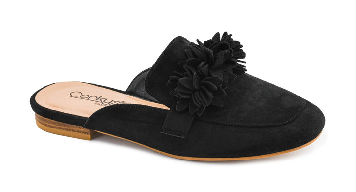 A black suede mule slipper with a large fabric flower embellishment on the strap, shown from a side angle against a white background. The insole is light tan with the brand name Corkus visible, and the outsole is light brown.