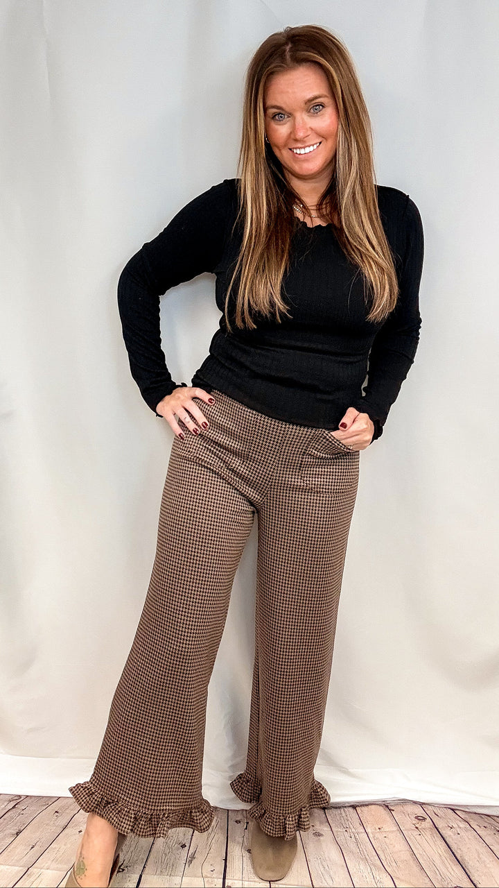 Woman wearing brown houndstooth pants with ruffle trim and black long sleeve top on plain background