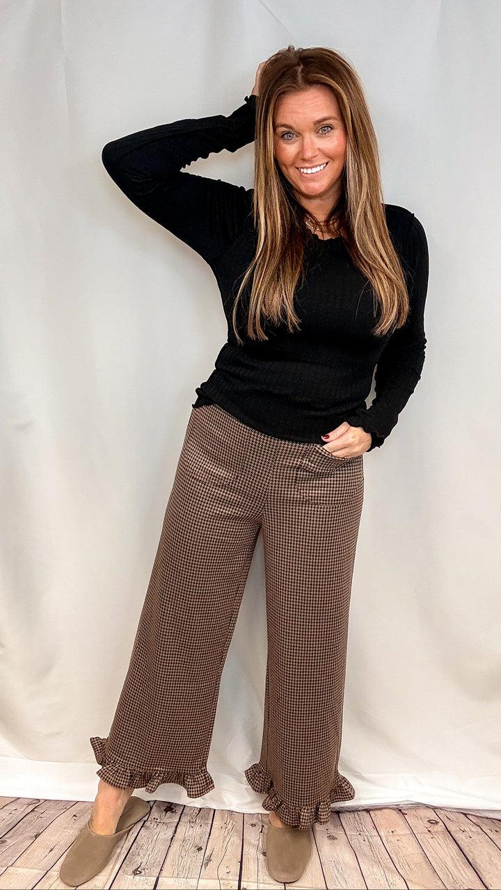 Woman wearing brown houndstooth pants with ruffle trim and black long sleeve top on plain background