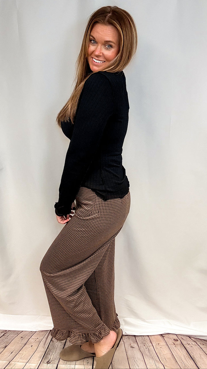 Woman wearing brown houndstooth pants with ruffle trim and black long sleeve top on plain background