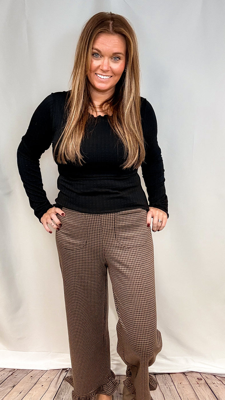 Woman wearing brown houndstooth pants with ruffle trim and black long sleeve top on plain background