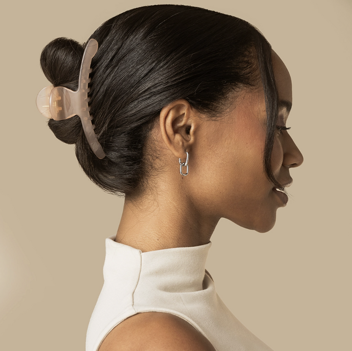 A woman in profile wearing a translucent beige claw-style hair clip that holds her dark hair in a low bun, paired with a white mock-neck top against a solid beige background.