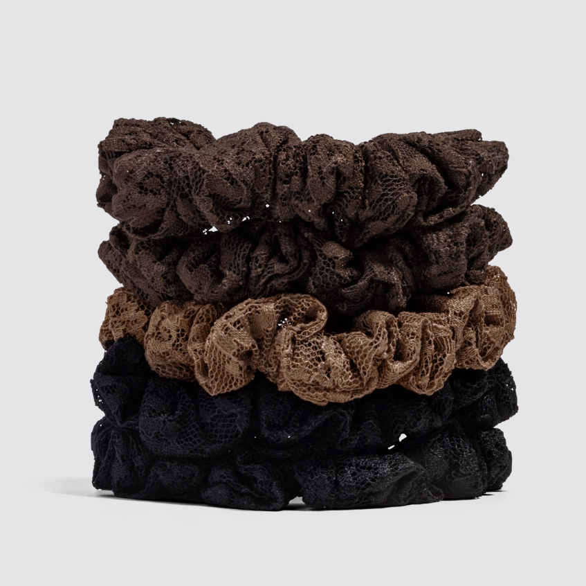 A stack of four textured lace scrunchies in dark brown, medium brown, black, and very dark brown, photographed on a light gray studio background.
