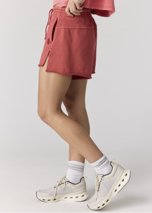 Woman wearing pink sweatshirt with red sweat shorts and tennis shoes on plain background