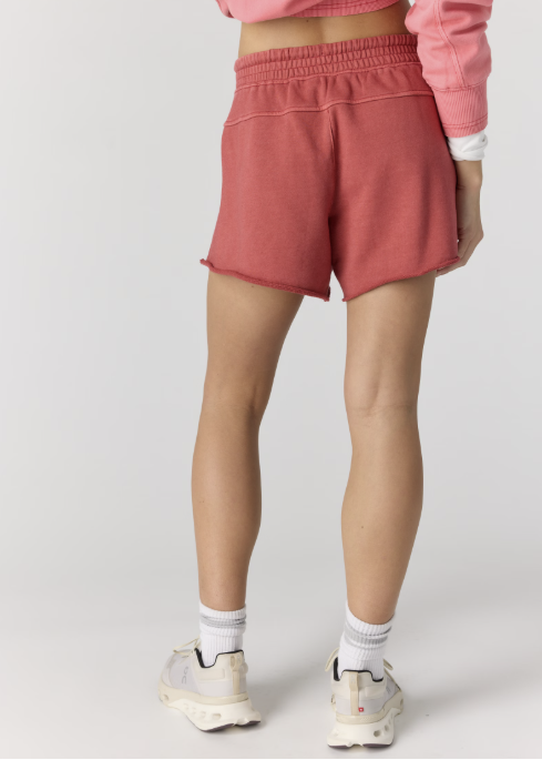 Woman shown from back wearing pink sweatshirt and red sweat shorts with tennis shoes on plain background