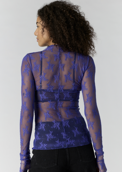 Woman shown from behind wearing blue lace top and bralette with black pants on plain background