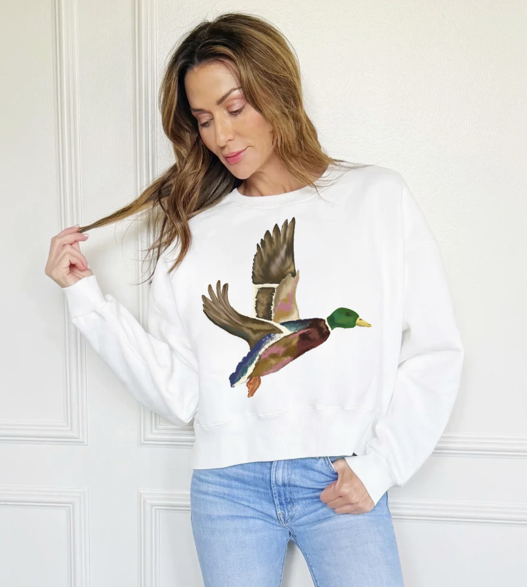 A woman wearing a white crewneck sweatshirt with a large, colorful graphic of a flying mallard duck on the front, paired with light blue jeans, standing against a white paneled wall.