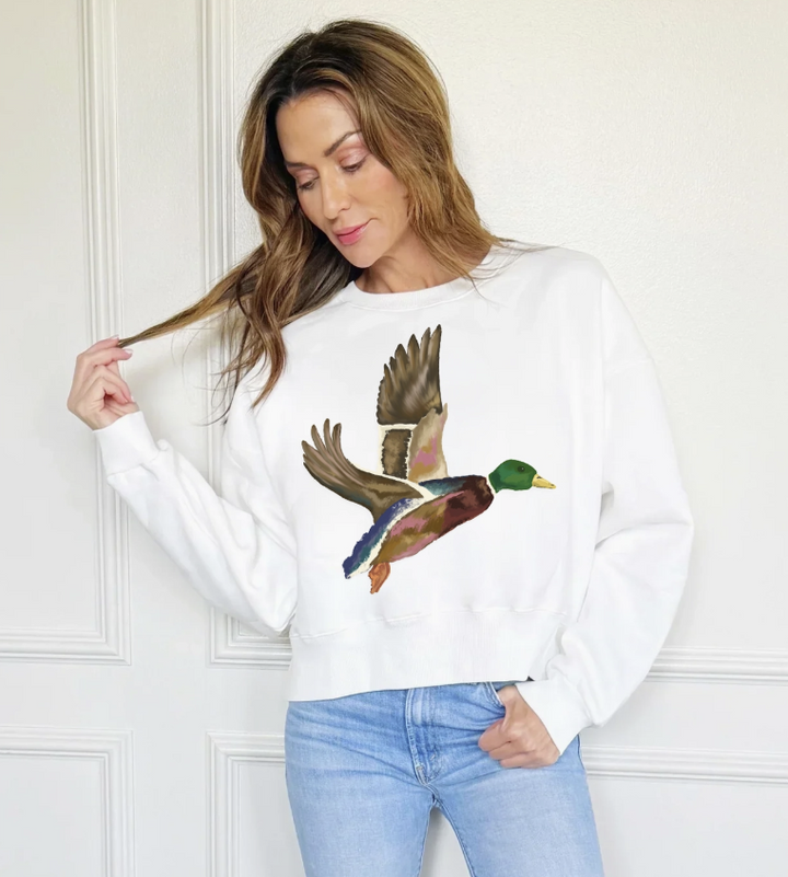 A woman wearing a white crewneck sweatshirt with a large, colorful graphic of a flying mallard duck on the front, paired with light blue jeans, standing against a white paneled wall.