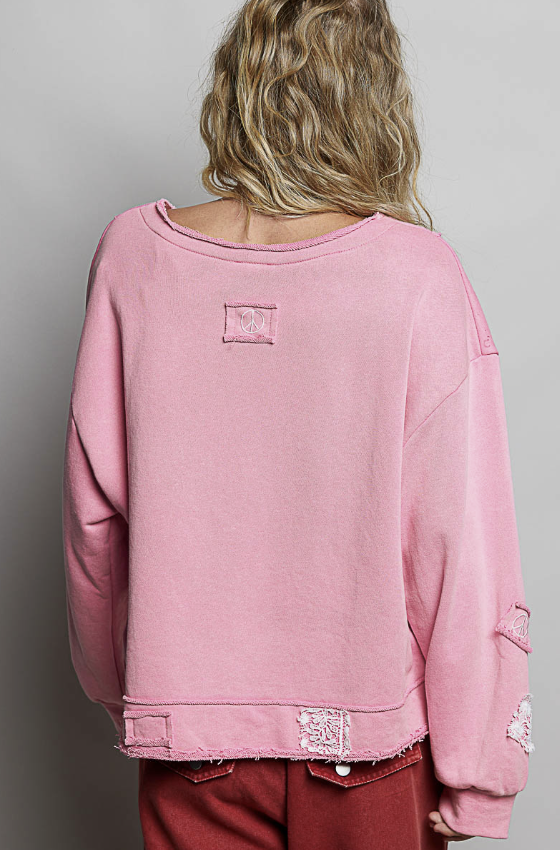 A person shown from the back wearing a pink oversized sweatshirt with distressed details, frayed hem, and a small square patch on the upper back, paired with dark red pants against a plain gray background.