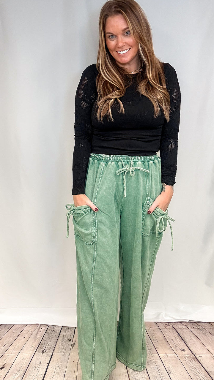 A woman modeling a black long-sleeve top and sage green wide-leg drawstring pants with side pockets, standing against a white studio backdrop with a light wood floor.