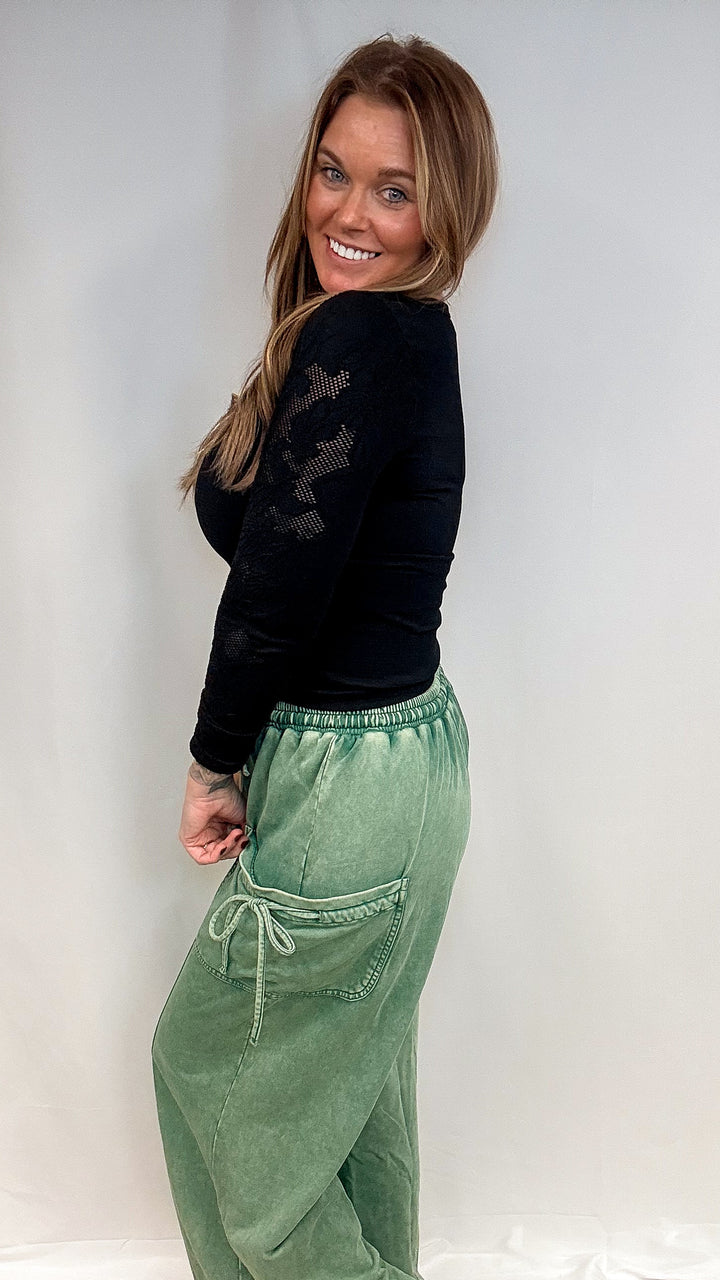 A woman modeling a pair of wide-leg sage green drawstring pants and a black long-sleeve top, standing against a white studio backdrop with a light wood floor.