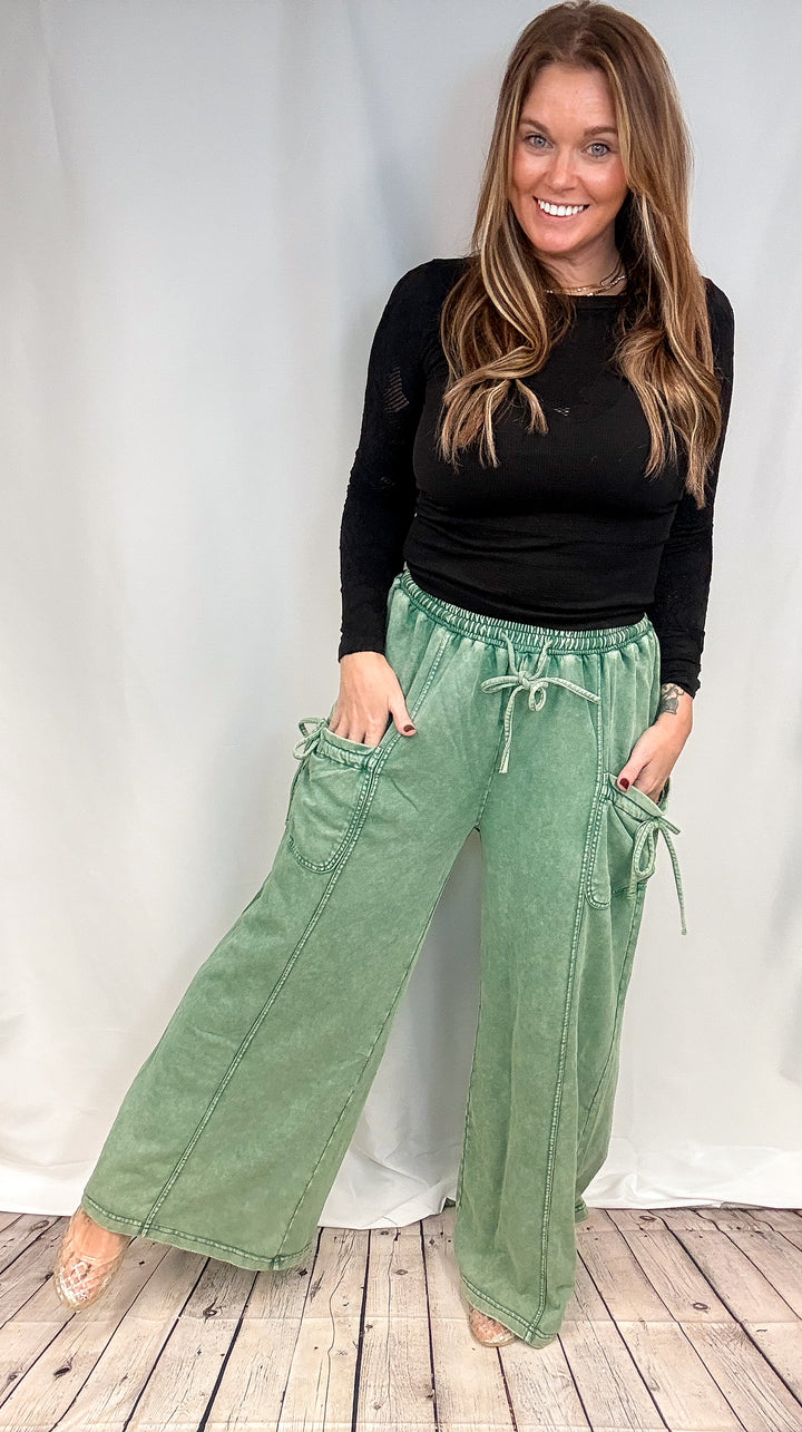 A woman modeling a pair of sage green wide-leg drawstring pants with side pockets, paired with a black long-sleeve top, standing against a white studio backdrop.