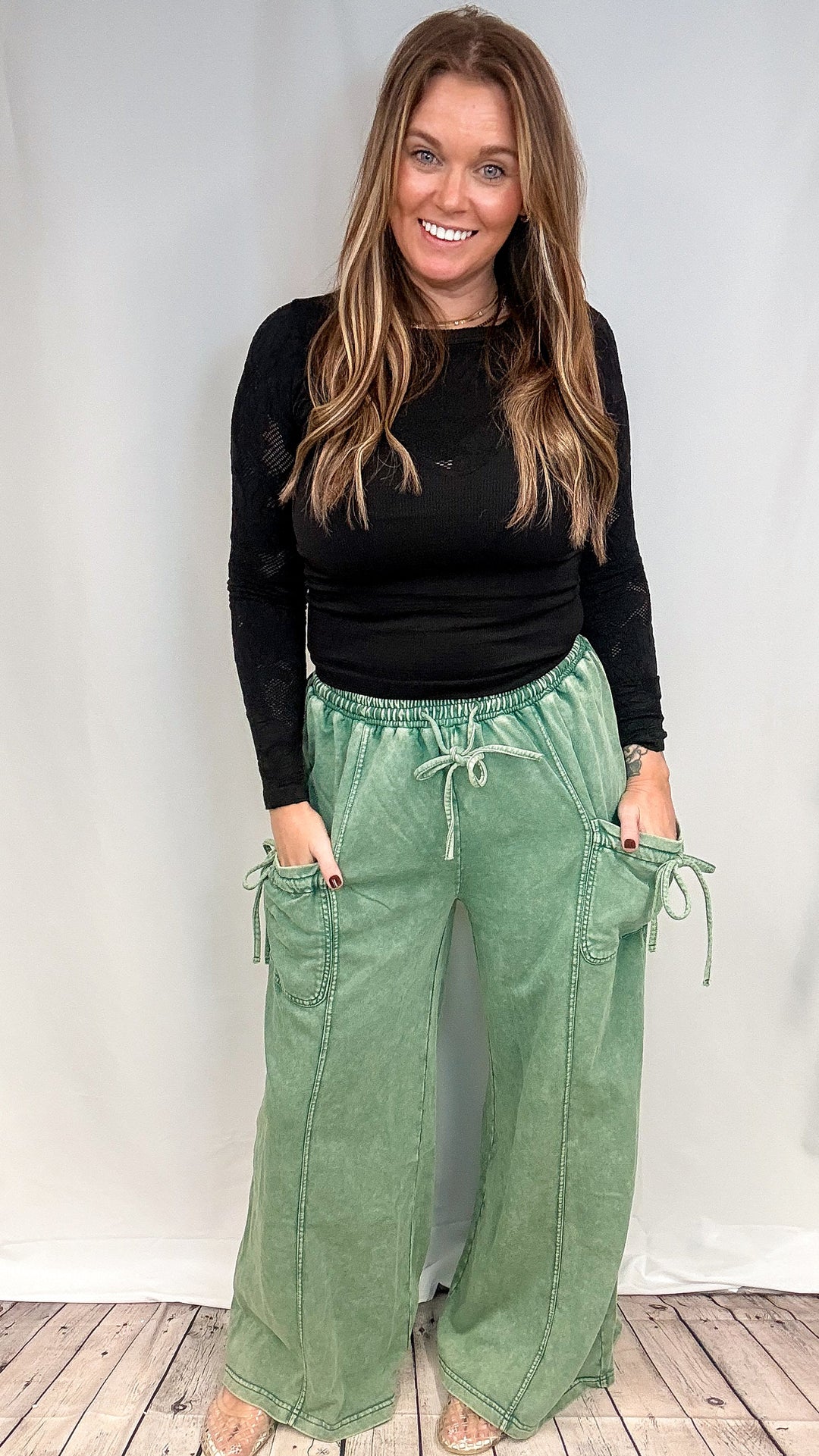 A woman modeling a black long-sleeve top and sage green wide-leg cargo pants with drawstrings, standing against a white studio backdrop on a light wood floor.