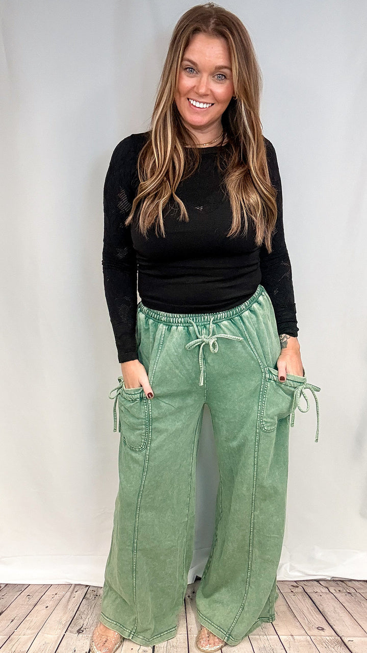 A woman modeling a black long-sleeve top and sage green wide-leg cargo pants with drawstrings, standing against a white studio backdrop on a light wood floor.