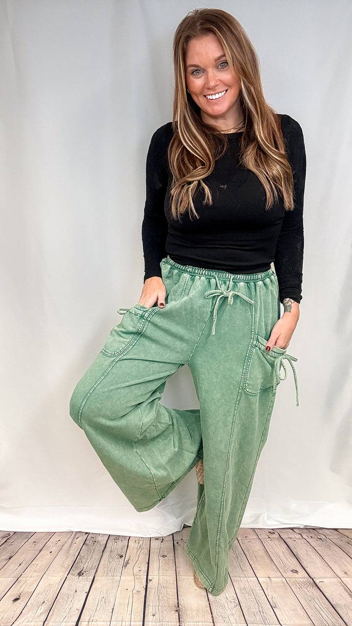 A woman modeling a pair of wide-leg sage green drawstring pants with a black long-sleeve top, standing against a white studio backdrop with a light wood floor.