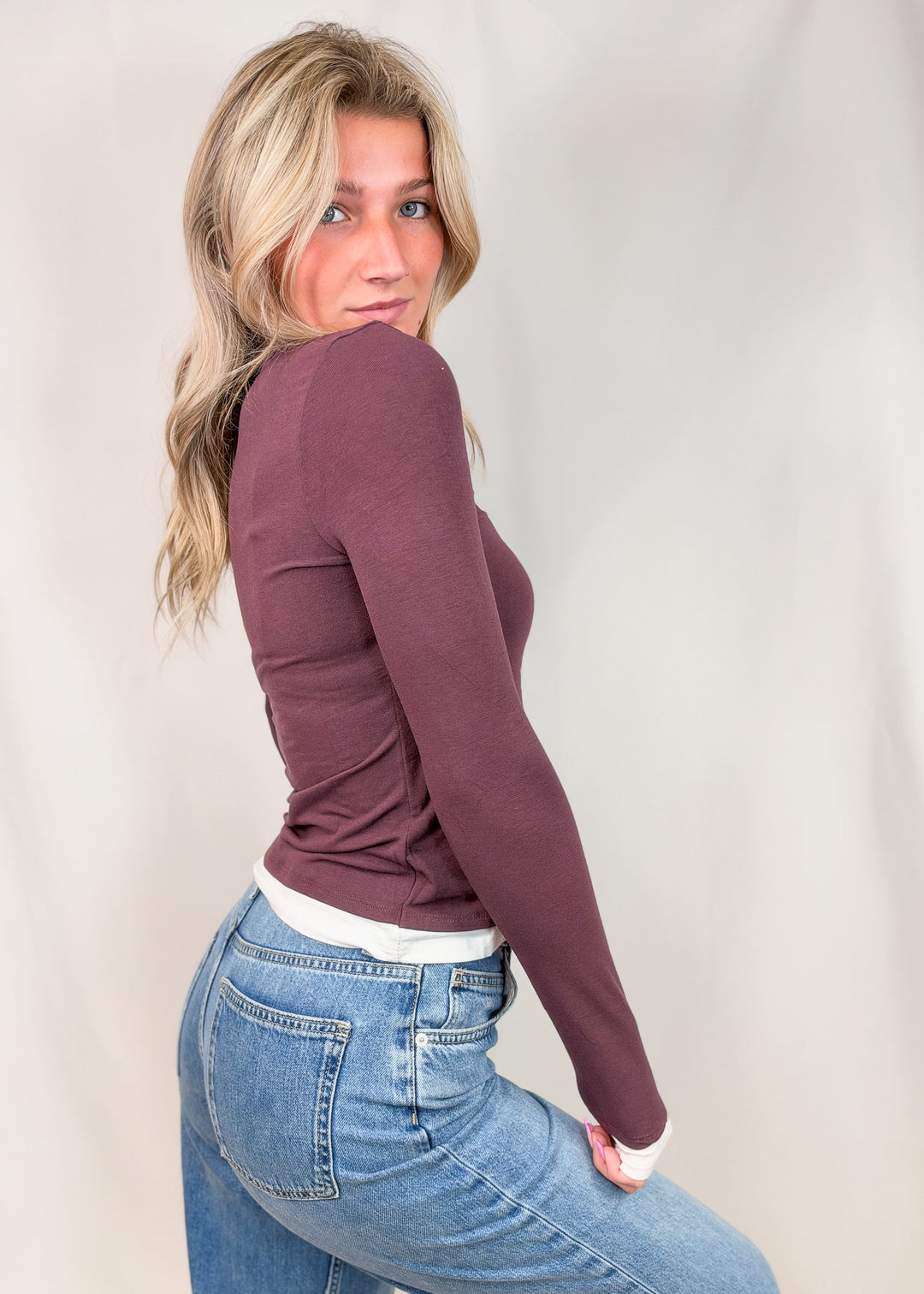 A woman posing in a studio wearing a fitted, long-sleeve crewneck top in a muted plum-brown color, tucked into light-wash blue jeans.
