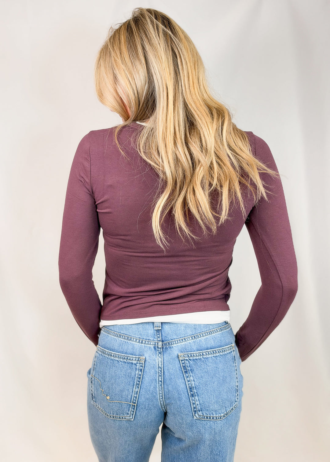 A woman viewed from behind wearing a fitted long-sleeve top in a muted plum-purple color and light-wash blue jeans against a plain light background.
