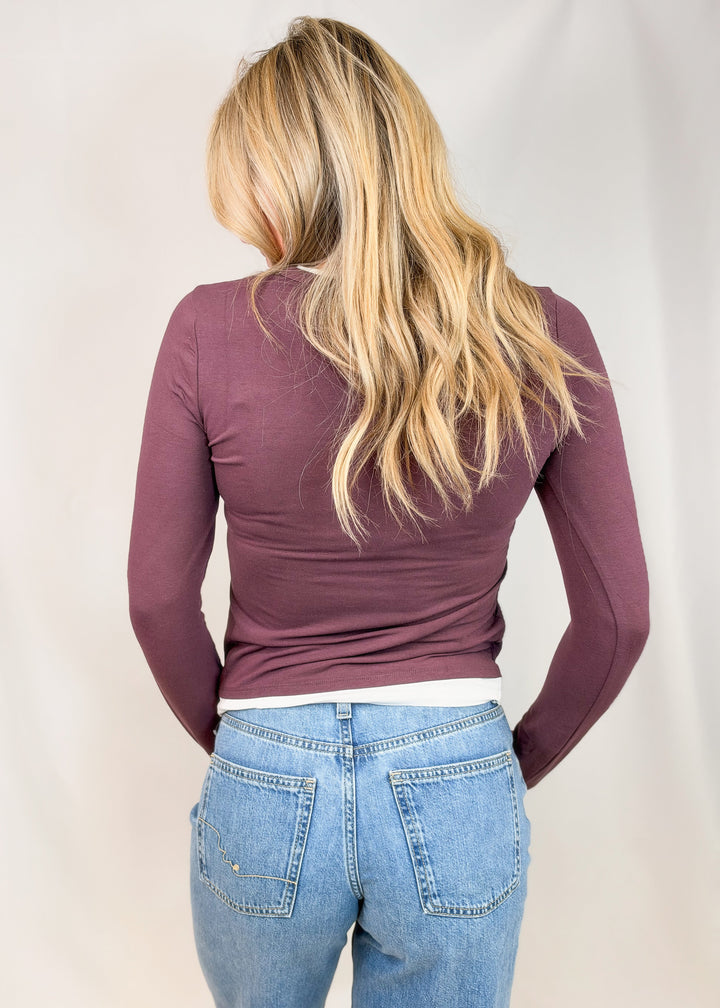 A woman viewed from behind wearing a fitted long-sleeve top in a muted plum-purple color and light-wash blue jeans against a plain light background.