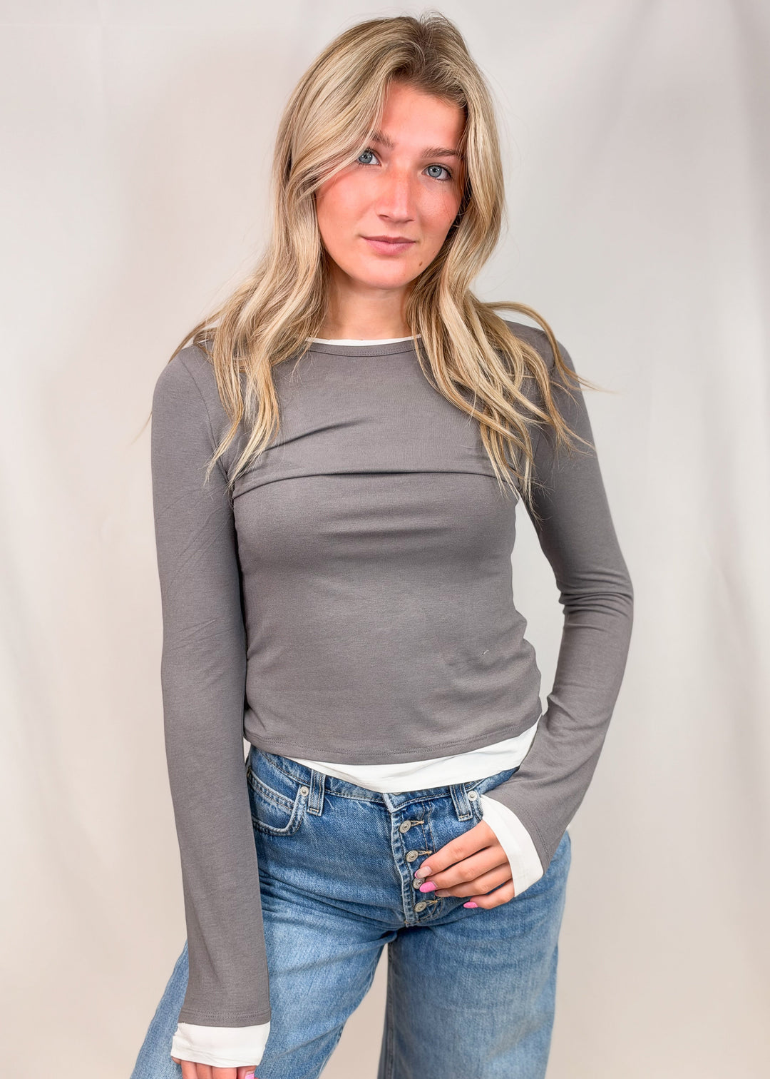 A woman modeling a fitted long-sleeve gray top with a white ribbed inner hem and white ribbed cuffs, paired with blue jeans, against a light neutral studio backdrop.