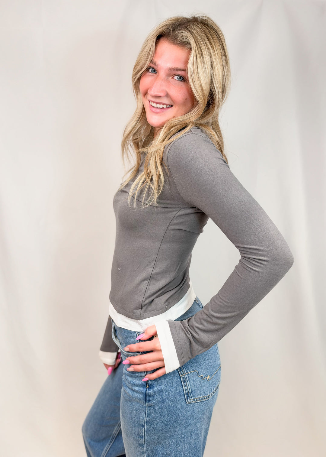 A smiling blonde woman posing in a fitted gray long-sleeve top with white cuffs and light blue jeans, photographed against a neutral draped studio backdrop.