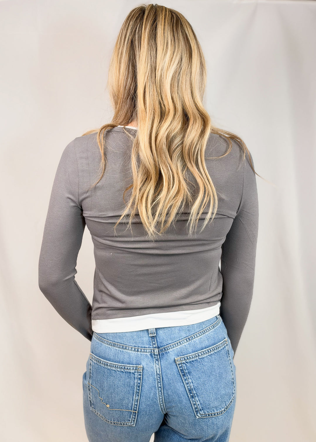 A woman viewed from behind wearing a fitted long-sleeve gray top and light blue jeans against a plain light backdrop.