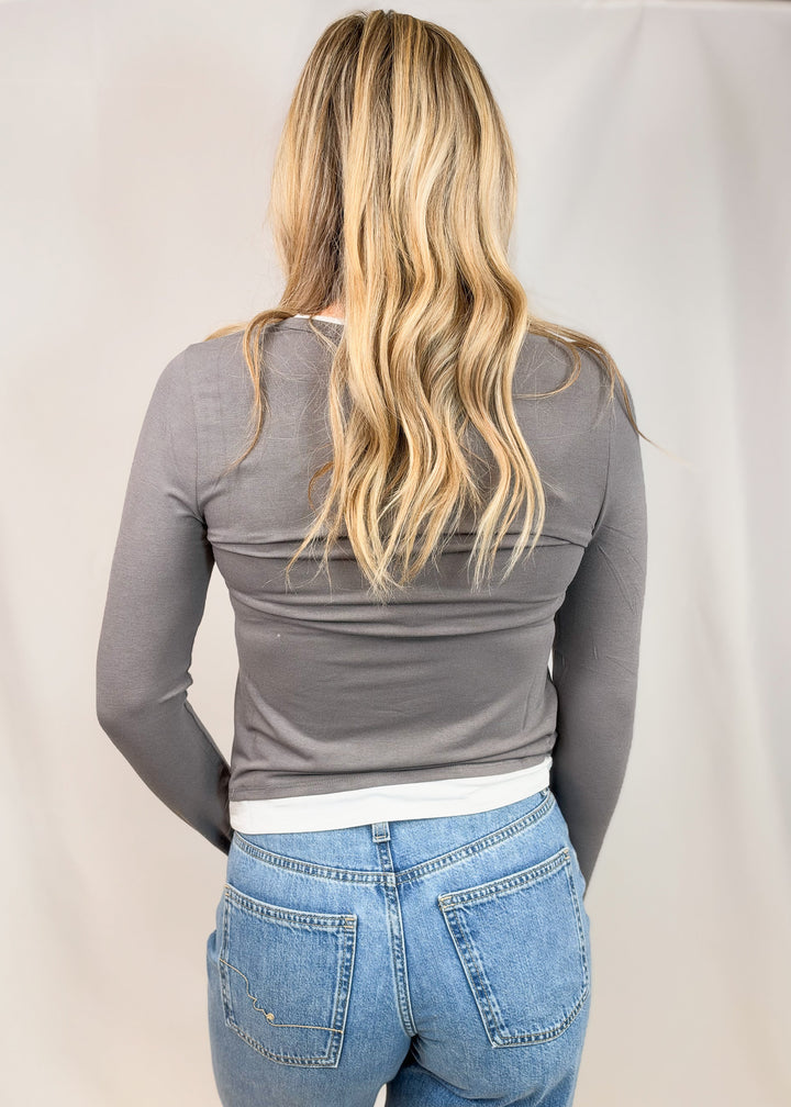A woman viewed from behind wearing a fitted long-sleeve gray top and light blue jeans against a plain light backdrop.