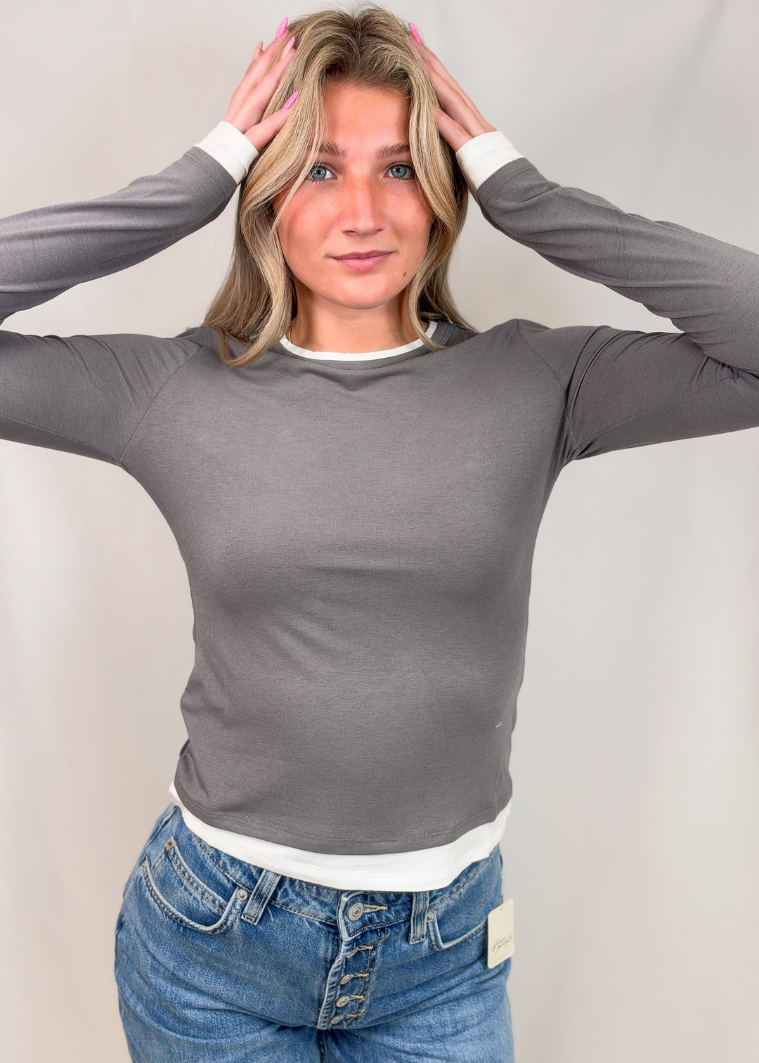 A woman wearing a fitted long-sleeve gray ribbed top with white trim at the collar and cuffs, paired with blue jeans, posing against a light neutral studio backdrop.