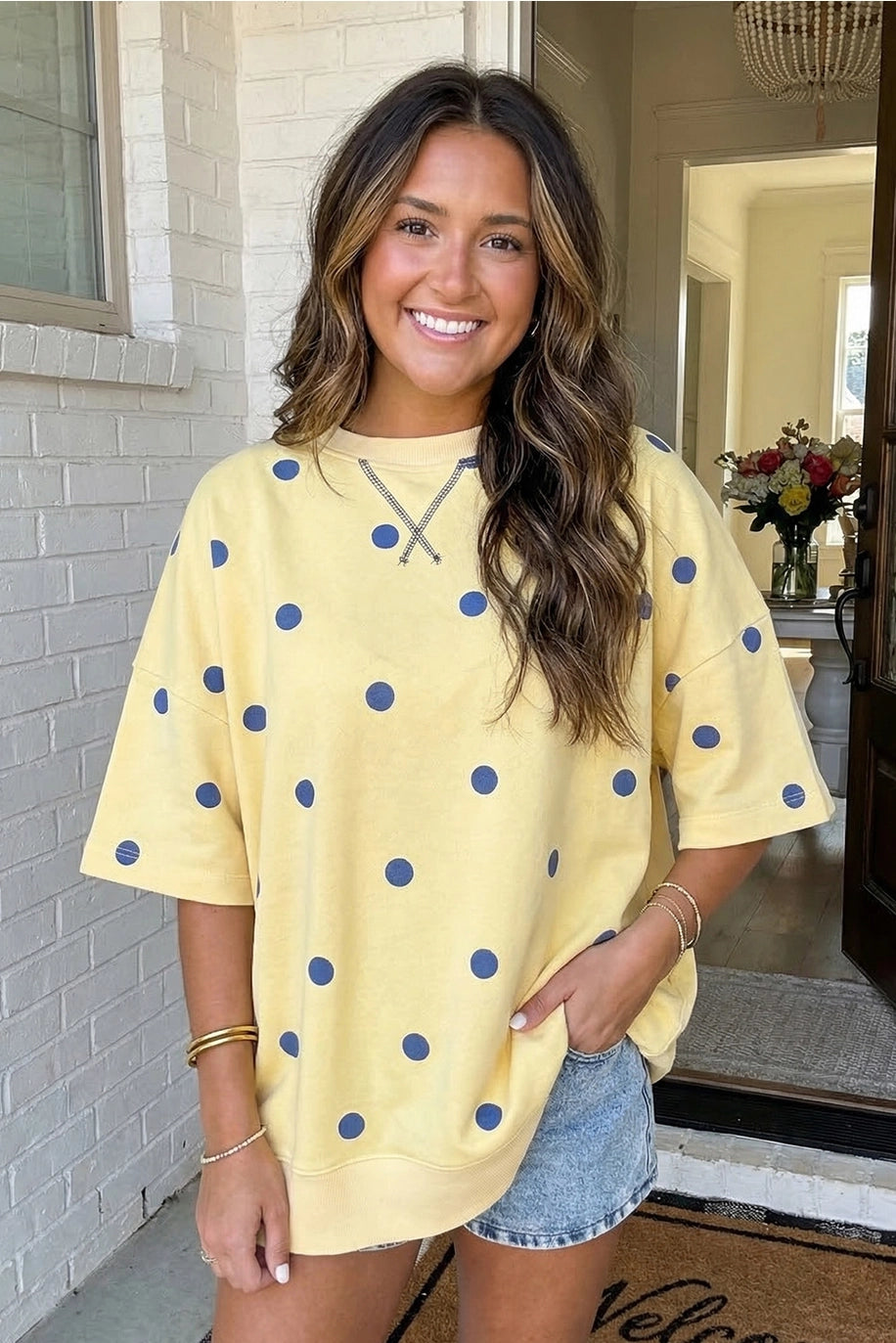 Woman wearing a yellow sweater with blue polka dots and denim shorts standing in a doorway.