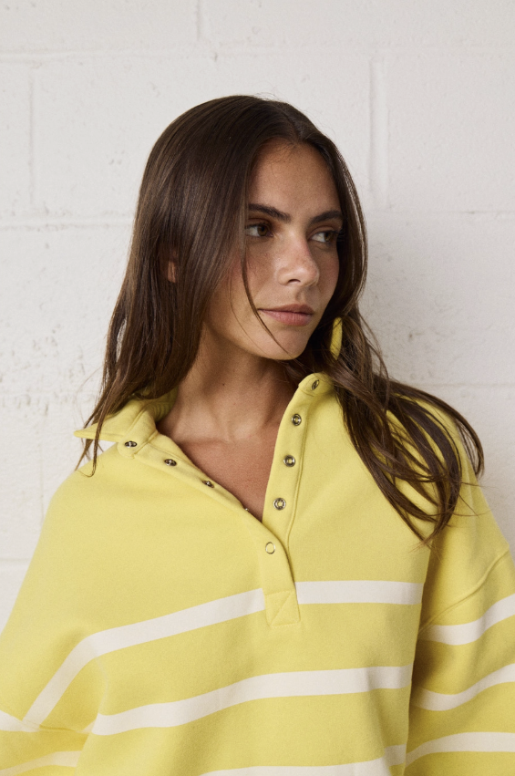A woman wearing a pale yellow pullover top with white horizontal stripes and a buttoned collar, photographed against a white painted brick wall.