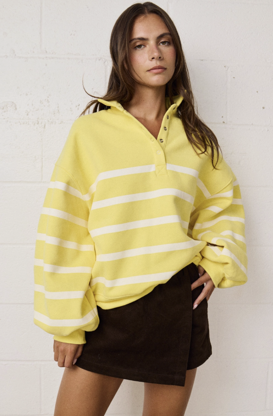 A woman wearing a bright yellow and white horizontally striped pullover sweatshirt with a buttoned collar, paired with a dark brown wrap mini skirt, standing against a white painted brick wall.