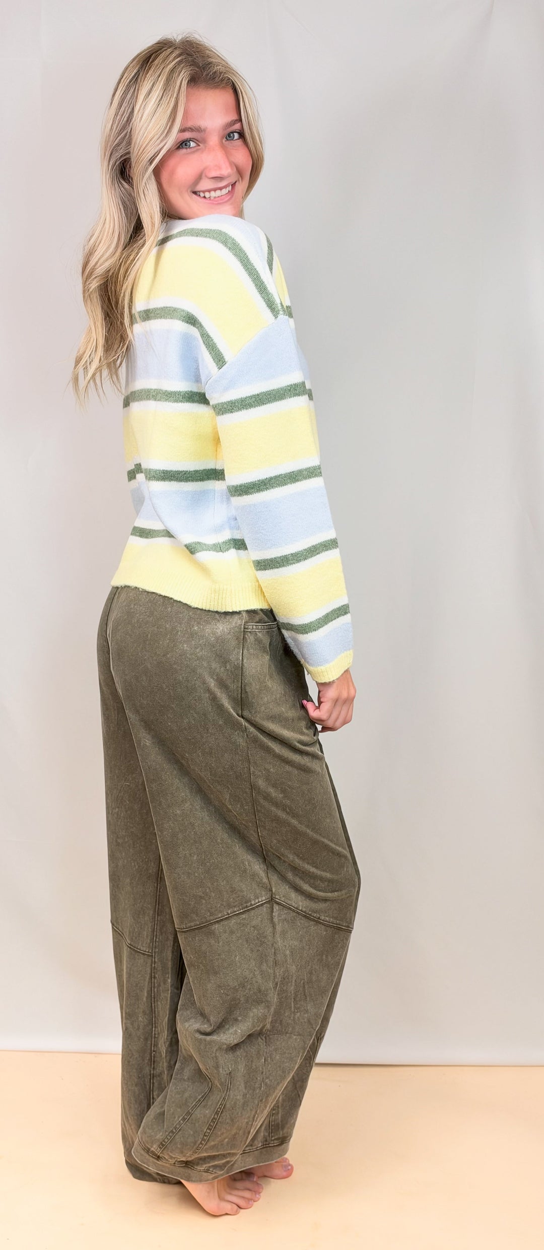 Woman wearing yellow, green and blue stripe cardigan with green pants on plain background
