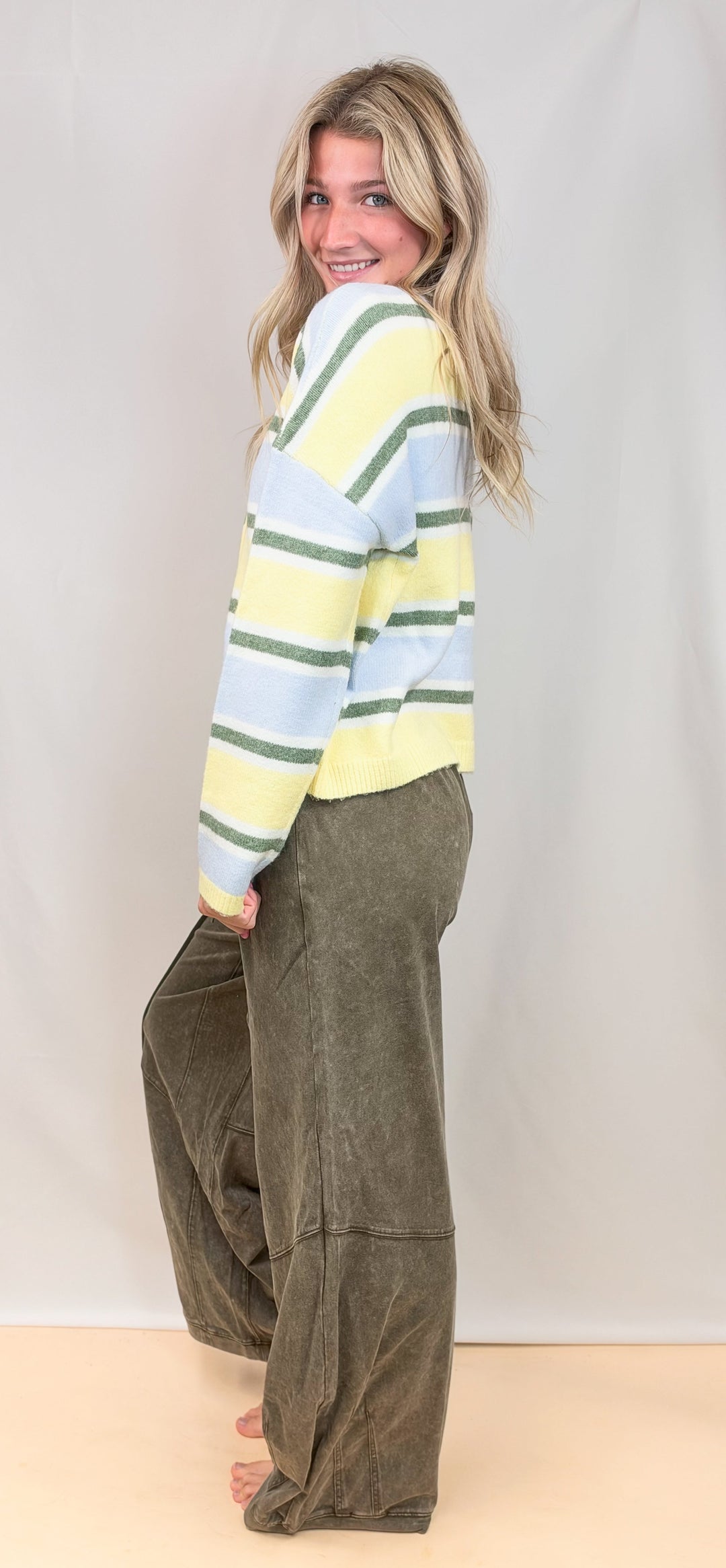Woman shown from side view wearing yellow, green and blue stripe cardigan with green pants on plain background