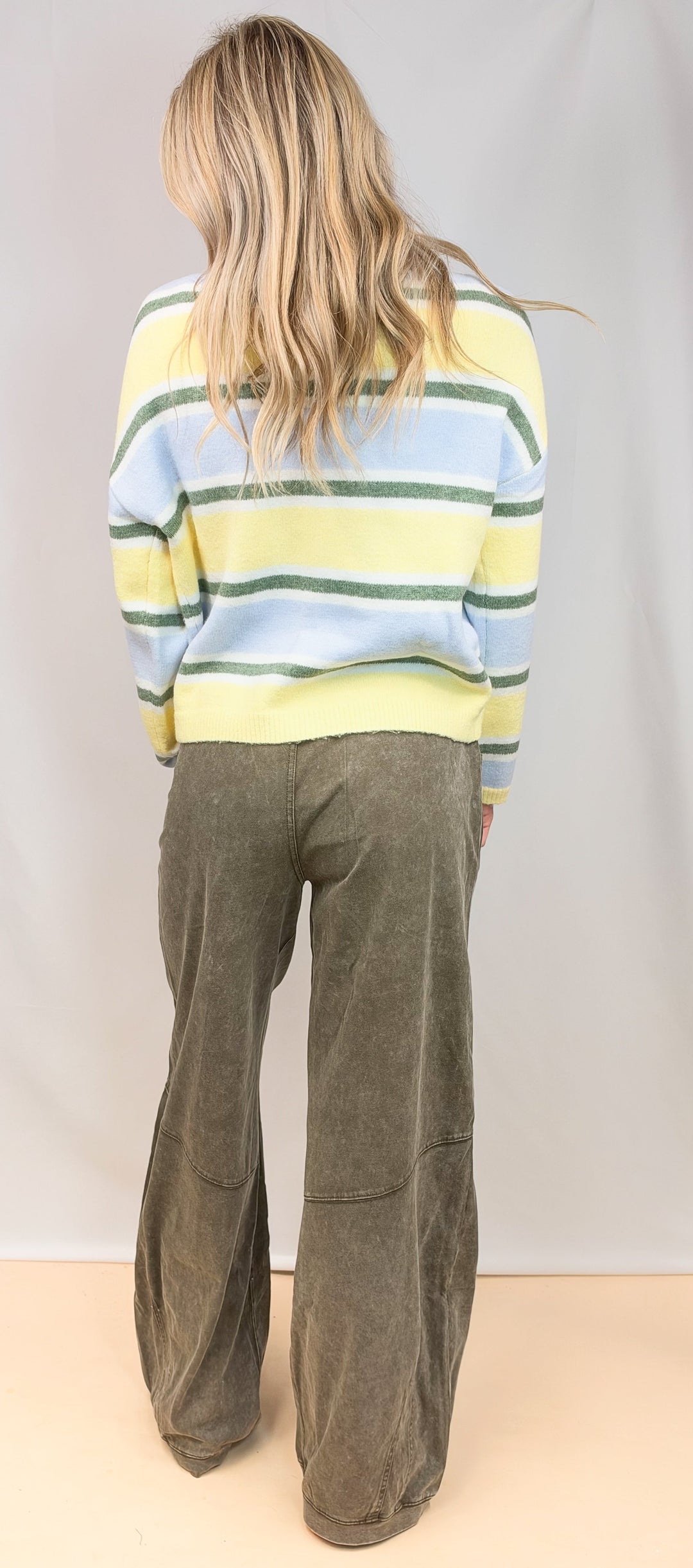 Woman shown from back wearing yellow, green and blue stripe cardigan with green pants on plain background