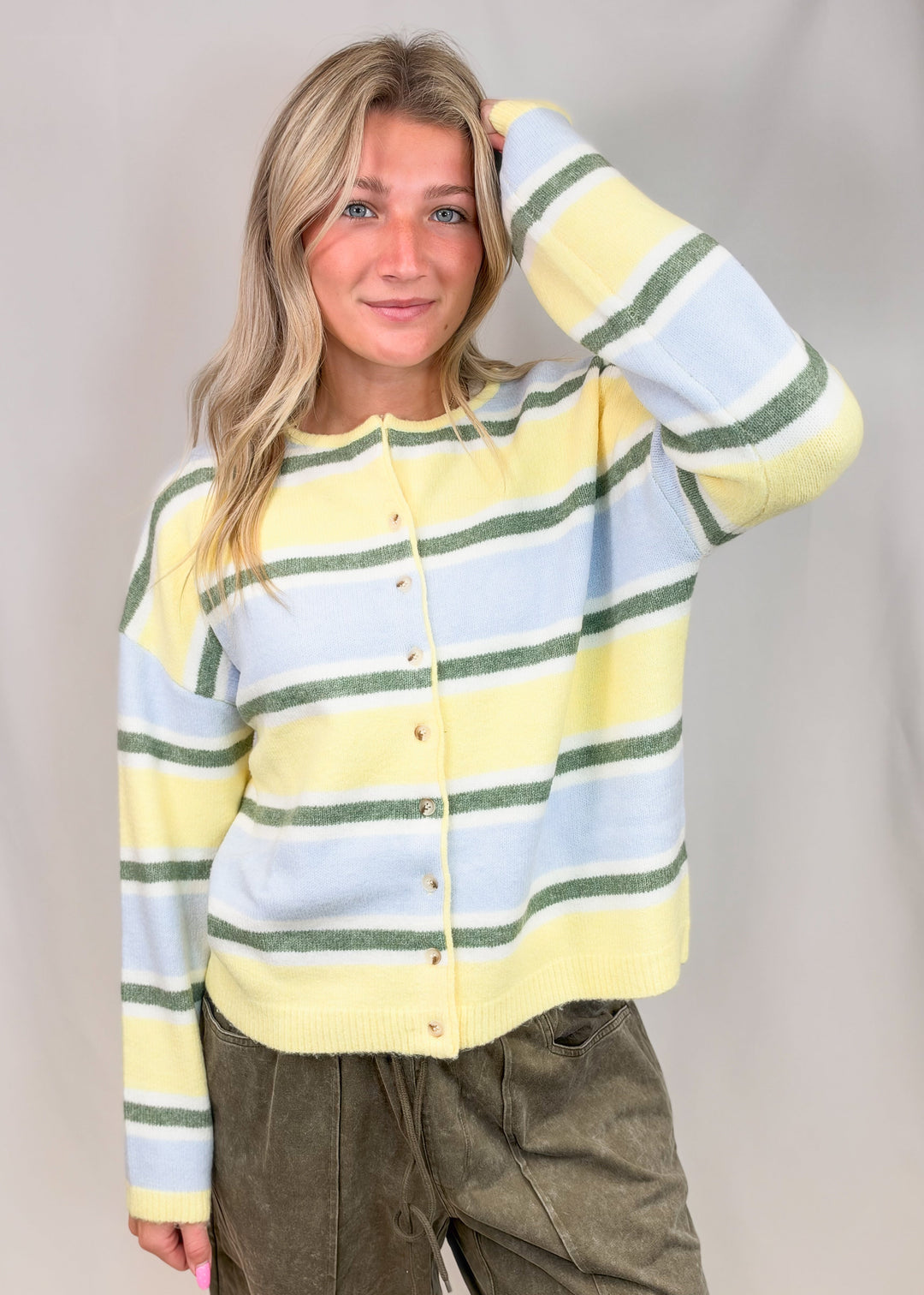 Woman wearing yellow, green and blue stripe cardigan with green pants on plain background