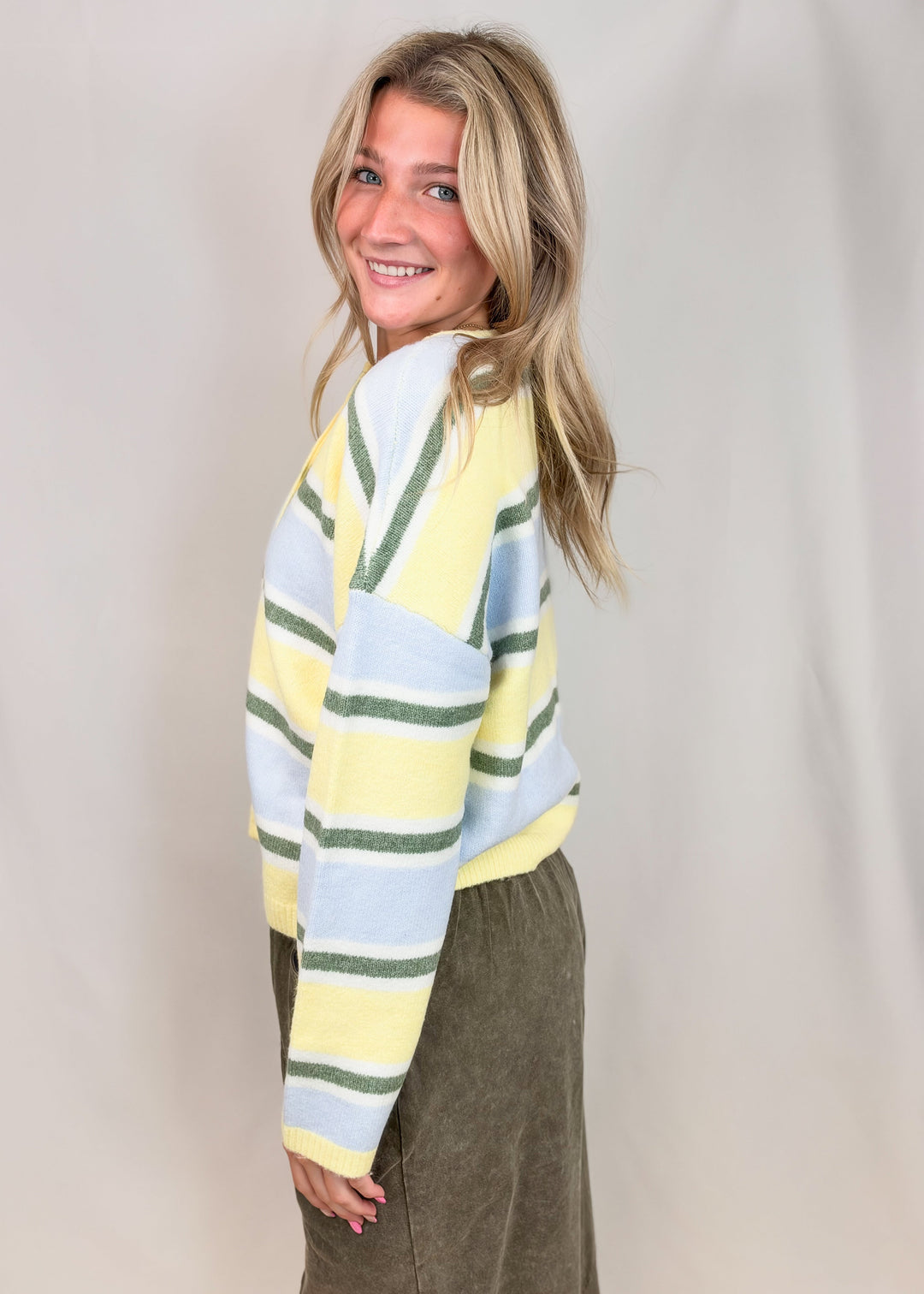 Woman shown from side view wearing yellow, green and blue stripe cardigan with green pants on plain background