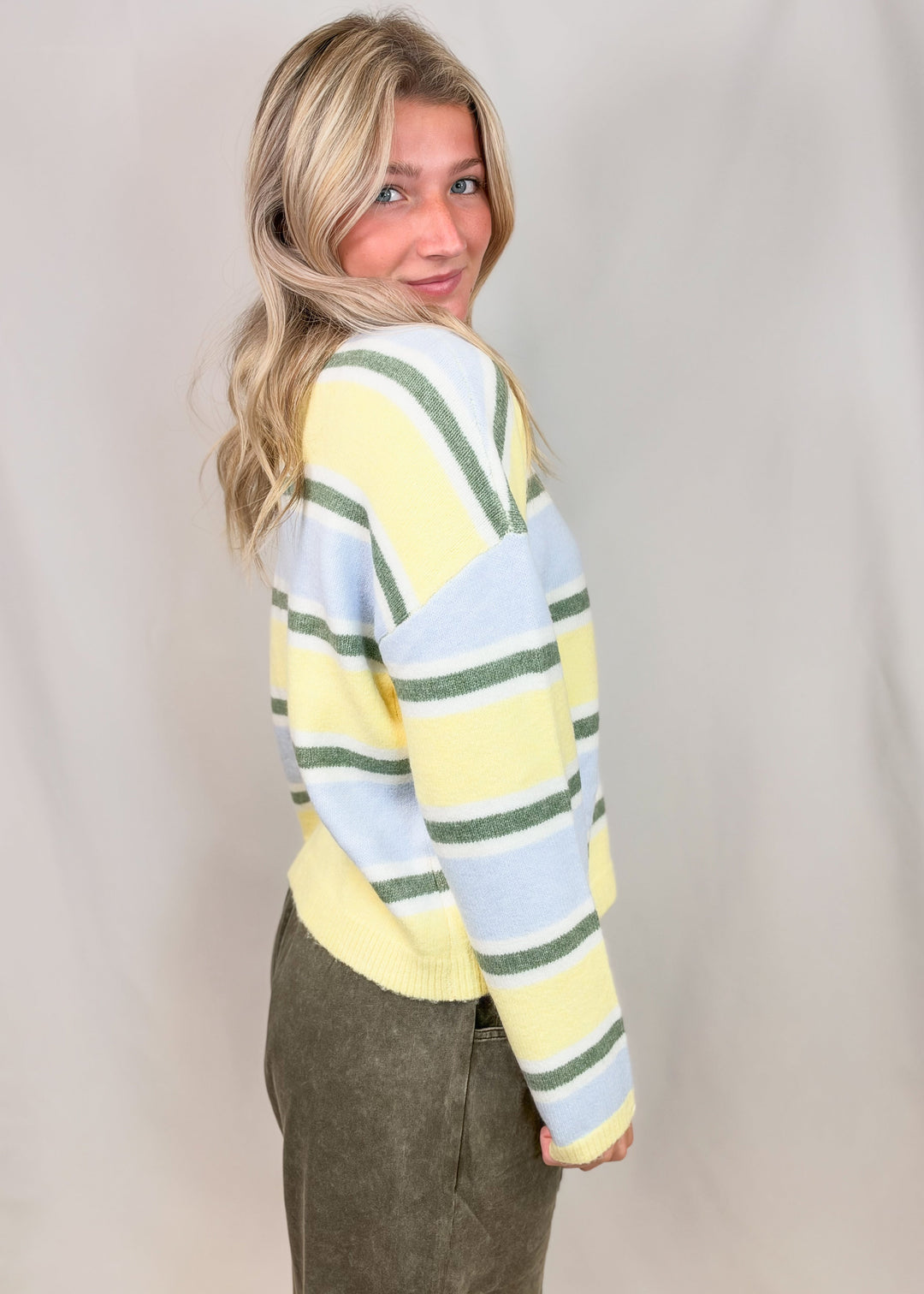 Woman shown from side view wearing yellow, green and blue stripe cardigan and green pants on plain background