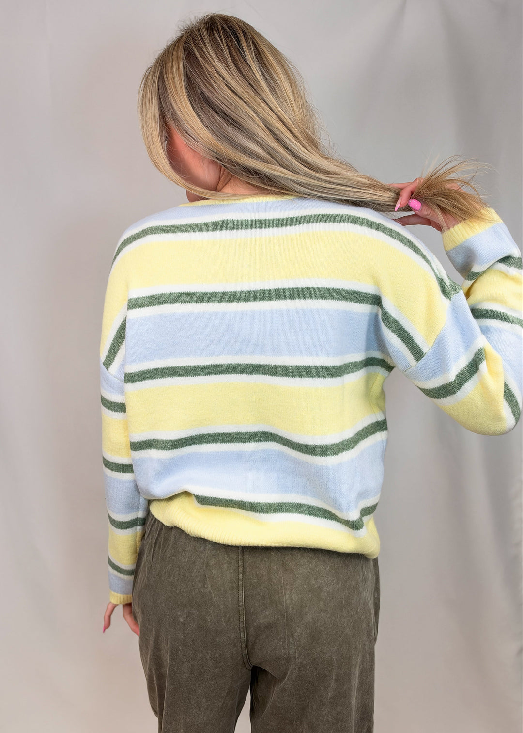 Woman shown from back view wearing yellow, green and blue stripe cardigan with green pants on plain background