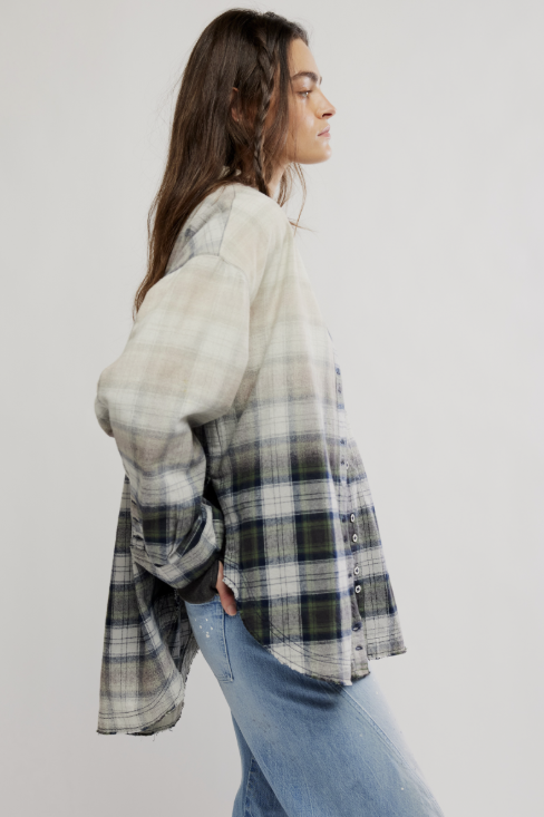 A woman in profile wearing an oversized plaid button-up shirt with a beige, gray, and green pattern, paired with light blue jeans, against a plain light gray studio background.
