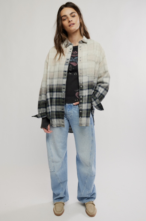 A model stands against a light gray studio background wearing a long-sleeve plaid overshirt with a black and white gradient fade, a black graphic t-shirt, light-wash blue jeans, and beige slip-on shoes.