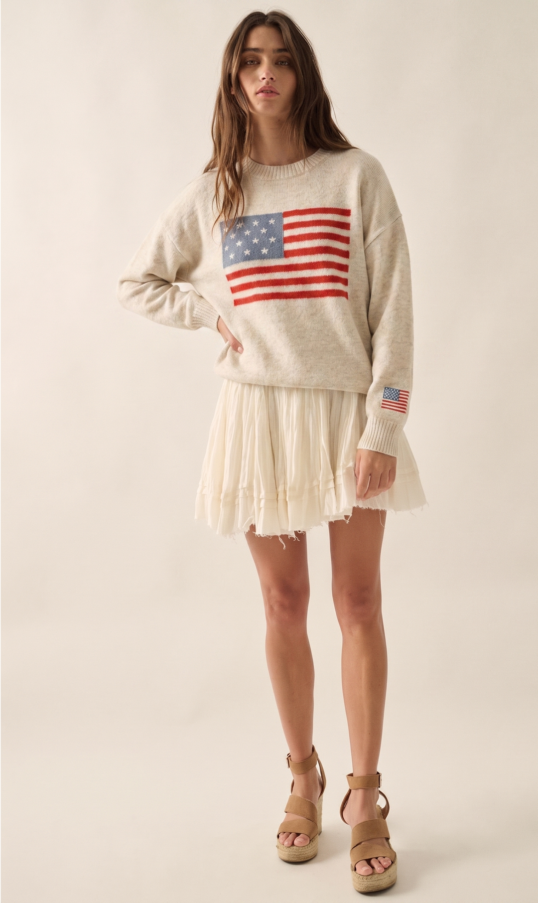 Woman wearing oatmeal colored sweater with American flag on front and sleeve against plain background