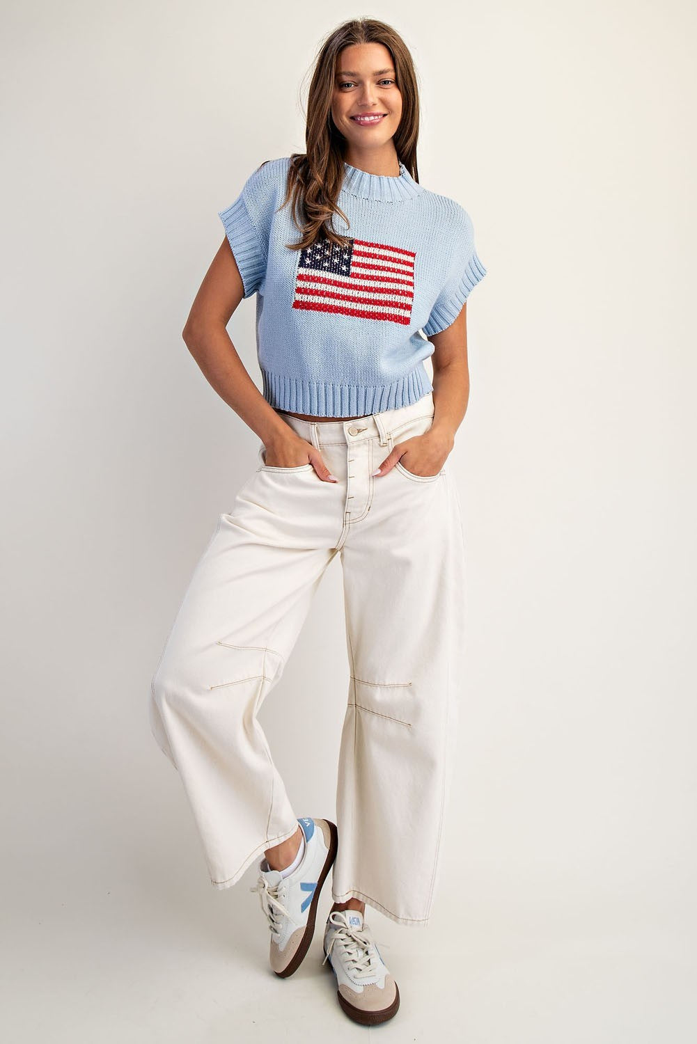 Woman wearing a light blue sweater with an American flag design and white pants on a plain background