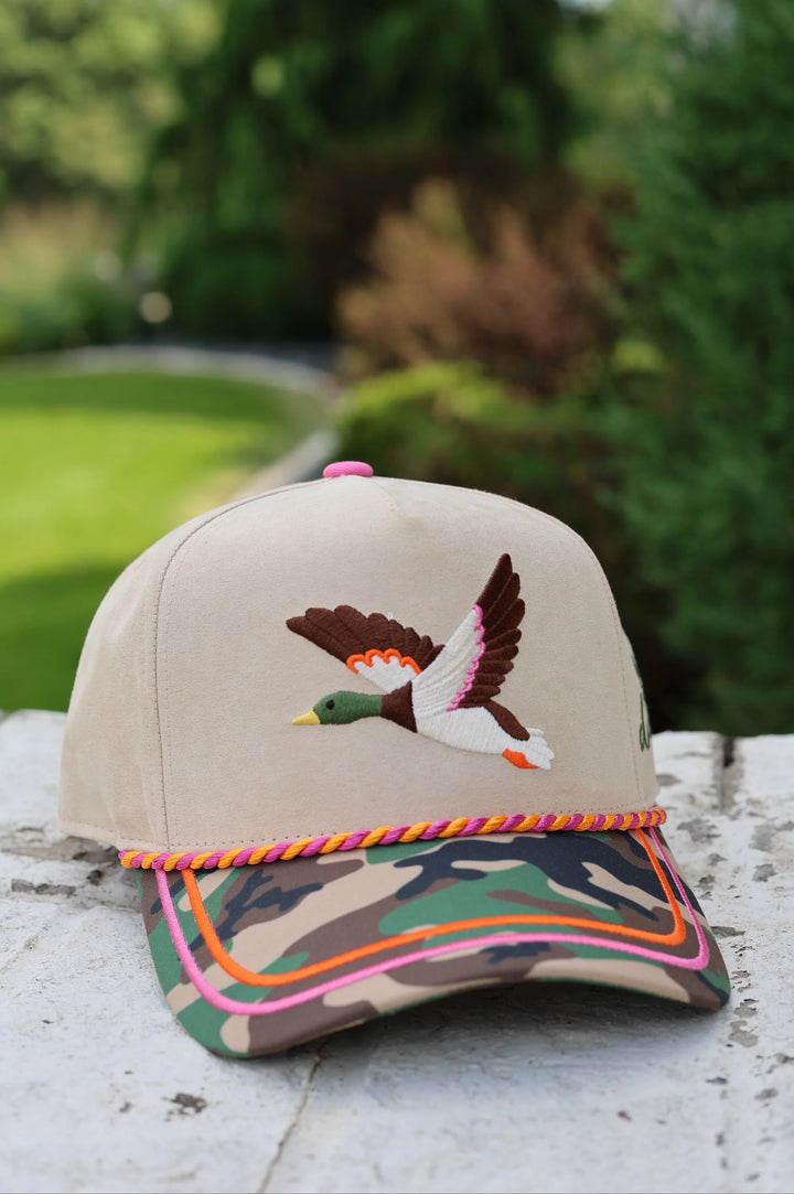 Beige trucker hat with camo bill, duck embroidery and pink and orange rope sitting outdoors
