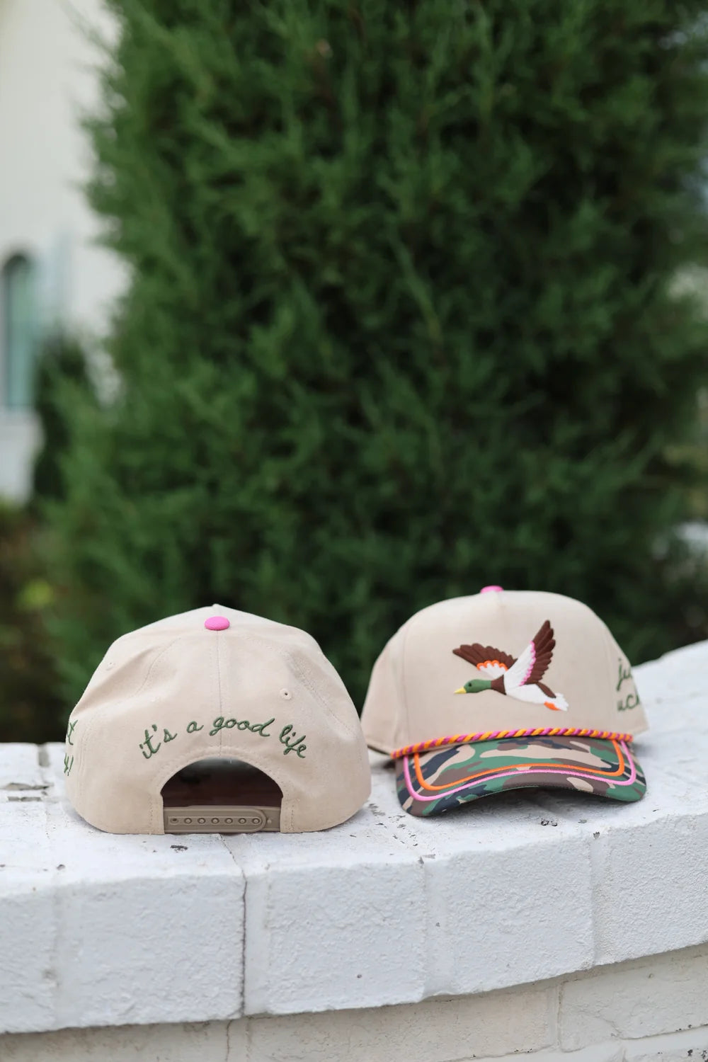 Two beige trucker hats with embroidered duck, it's a good life on back, camo bill and orange and pink rope sitting outdoors