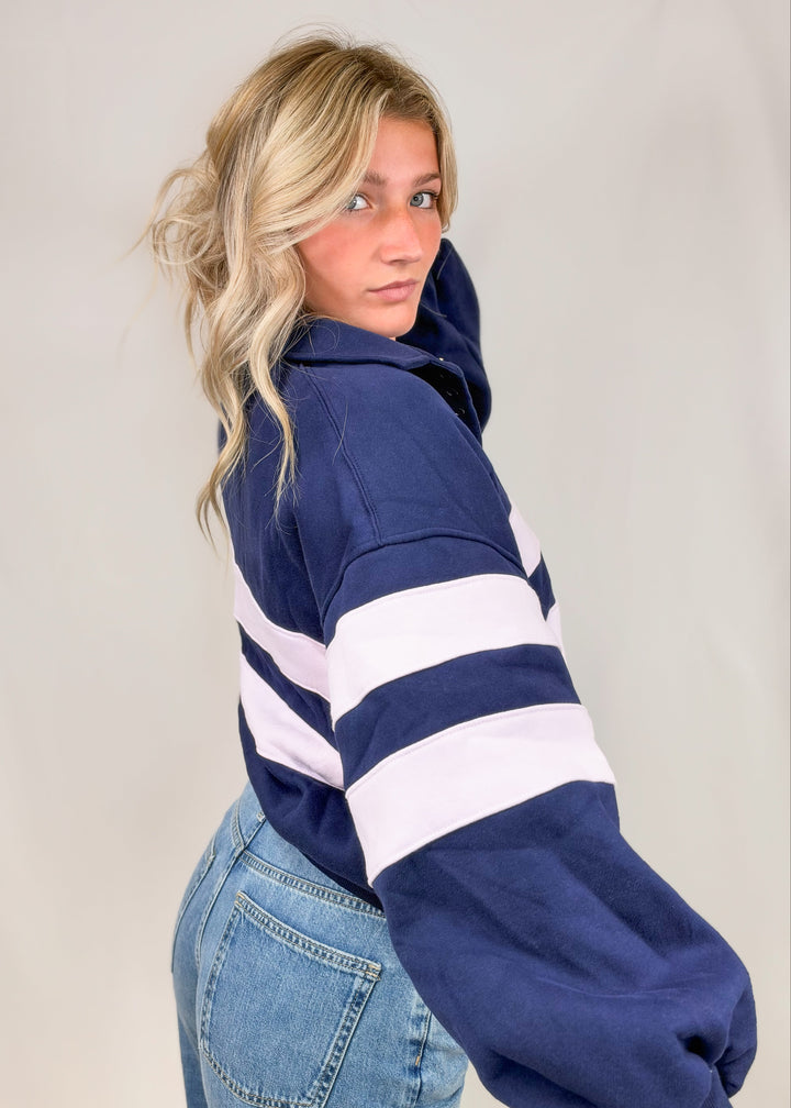 Woman shown from side view wearing navy sweatshirt with lilac stripes and jeans on plain background