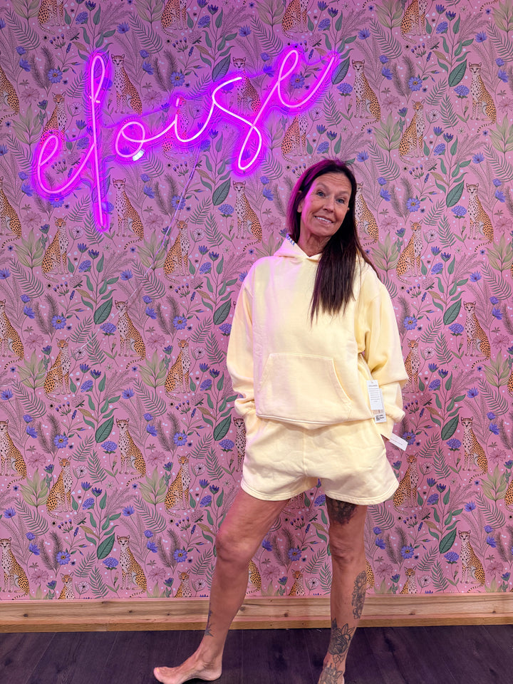 Woman wearing matching light yellow sweat shorts and sweatshirt on pink floral background