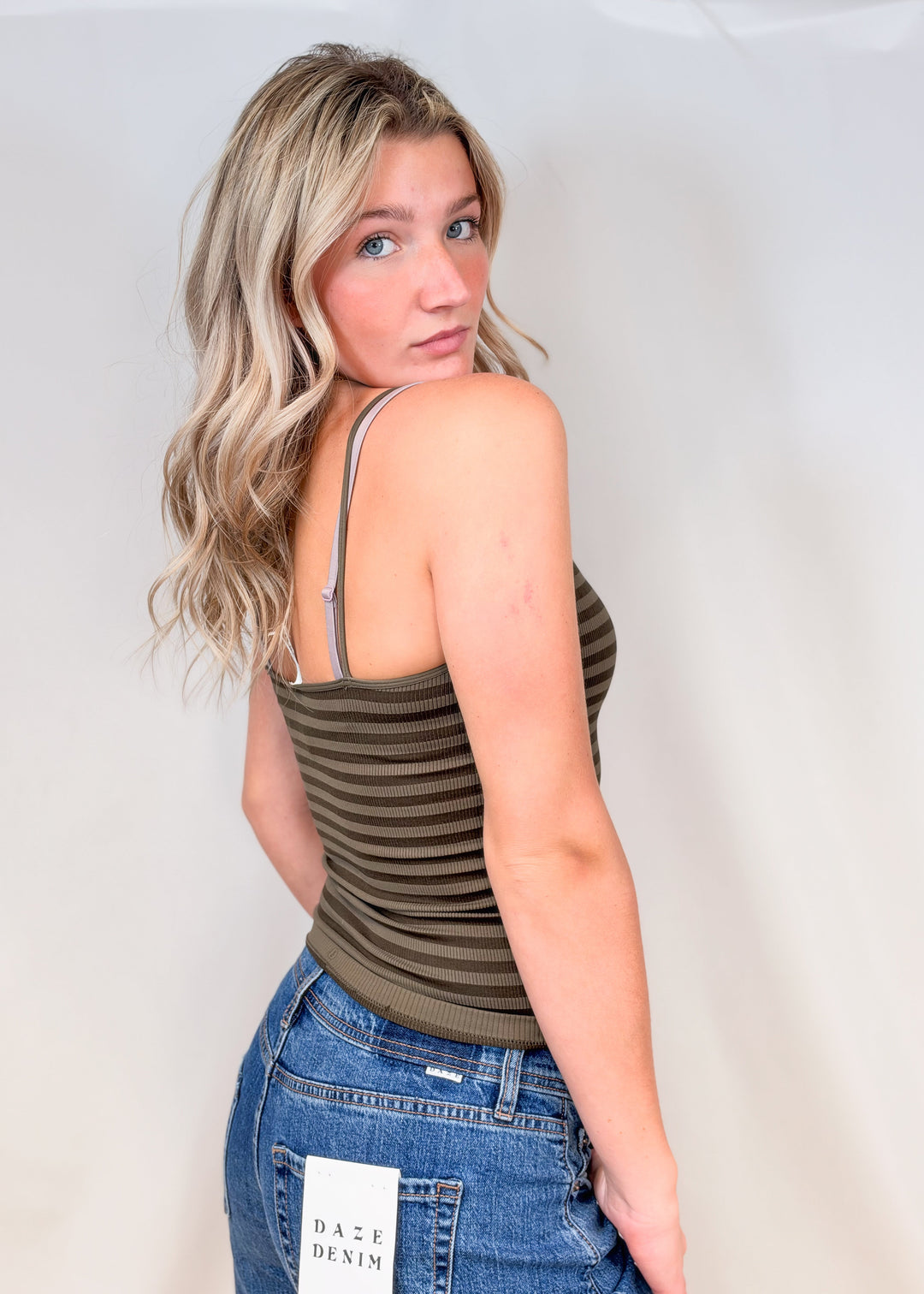 Woman shown from side view wearing green stripe tank and jeans on plain background