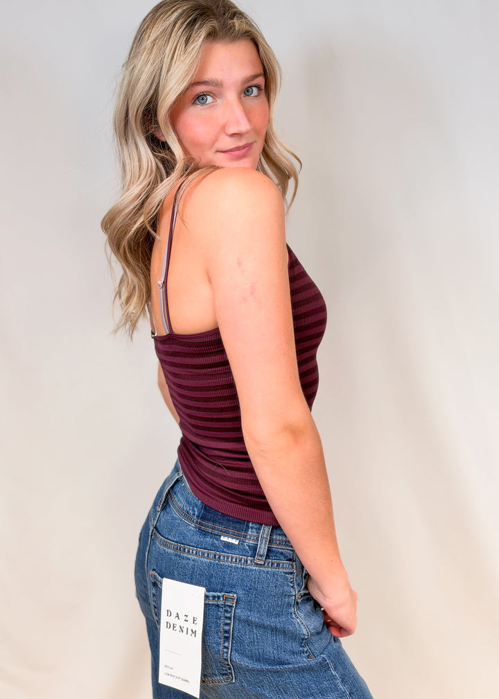 Woman shown from side view wearing burgundy stripe tank and jeans on plain background