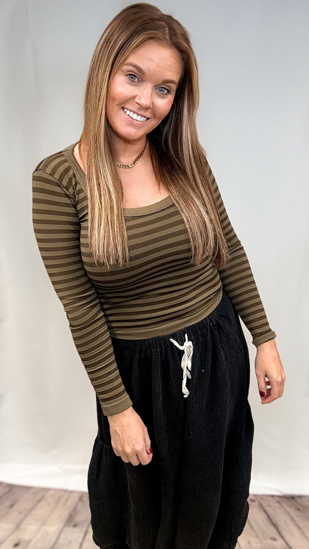 Woman wearing olive green stripe long sleeve top and black pants on plain background